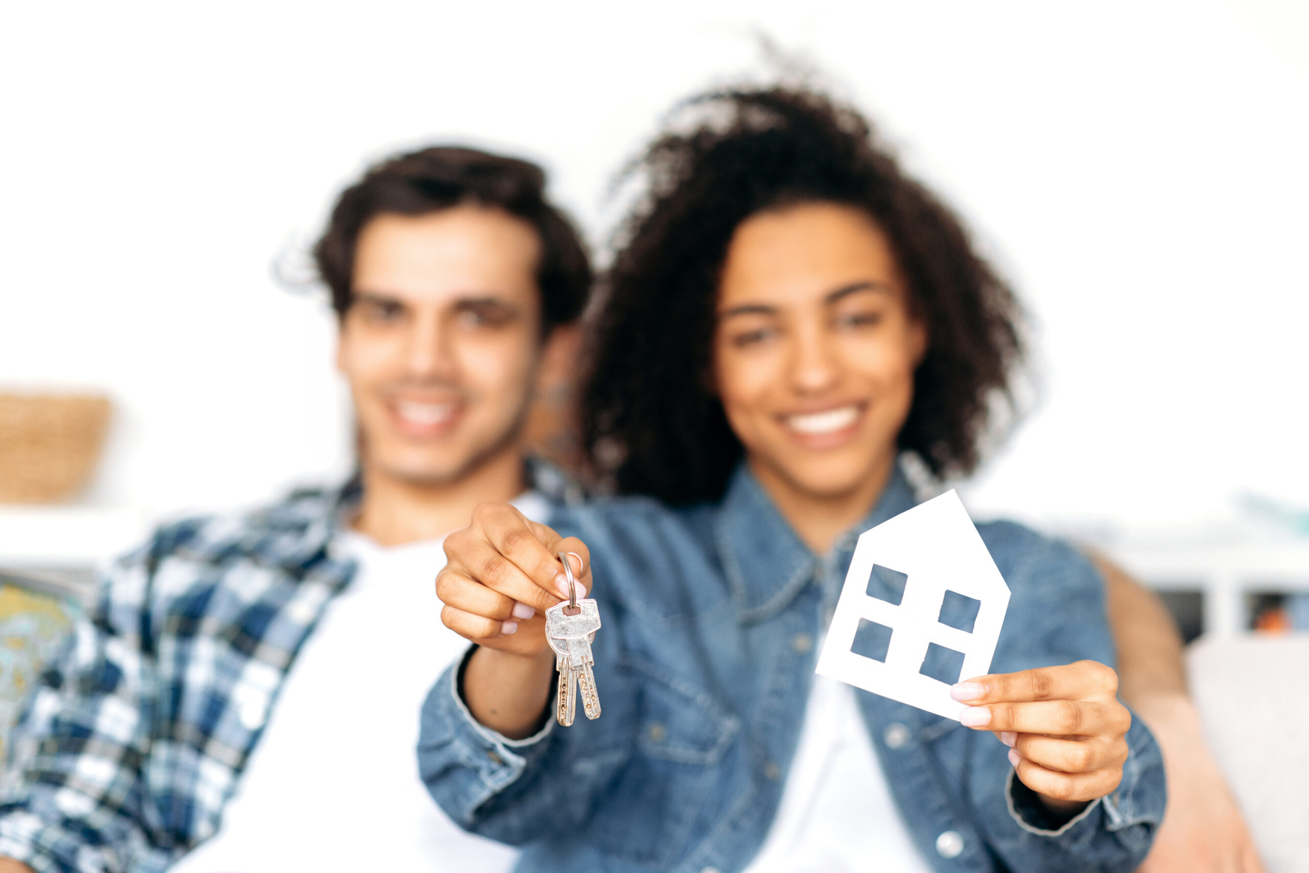 Joyful happy multiethnic young married couple is buying property. Blurred african american girl holding a cardboard house and keys while sitting on the couch with her hispanic boyfriend at new home
