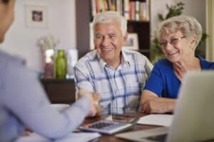 An older man and woman sit at a desk, smiling and shaking hands with another person—reviewing papers, a tablet, and calculator as they discuss using a Reverse Mortgage to Fund Your Retirement.