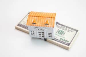 A small model house placed on top of two stacks of hundred dollar bills on a white background, illustrating how a reverse mortgage can fund your retirement.
