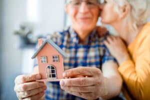 An older man holds a small model house while a woman leans in and kisses him on the cheek, celebrating their decision to use a Reverse Mortgage to Fund Your Retirement.