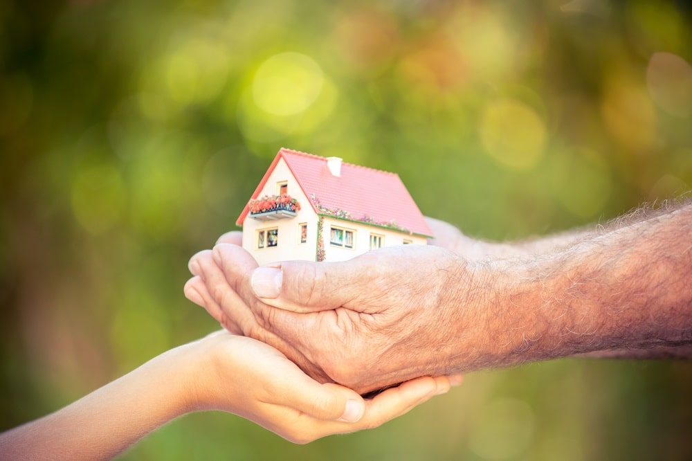 An elderly hand and a young hand hold a small model house together, symbolizing the transfer or sharing of home ownership—an idea echoed by using a Reverse Mortgage to Fund Your Retirement.