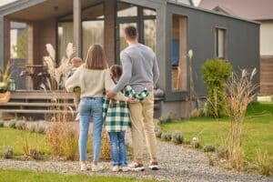 A family of four, seen from behind, stands together on a stone path in front of a modern house with a garden—an inspiring scene for those exploring FHA Loans for Low Credit Scores to achieve their homeownership dreams.
