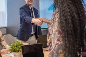 Two people in business attire shake hands across a table in an office meeting room, discussing DSCR Loans for Property Investors, with laptops and a plant visible on the table.
