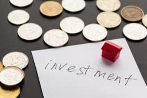 Coins scattered on a dark surface, a small red toy house, and a piece of paper labeled "investment" in black ink symbolize DSCR Loans for Property Investors.