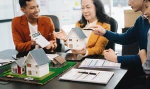 Four people in an office discuss real estate, holding model houses and a calculator, with documents and miniature houses on the table as they explore FHA Loans for Low Credit Scores.