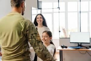 A man in military uniform is greeted by a smiling woman and girl in an office-like room with a desk and computer, perhaps learning how VA Loans work to support their family's next step.