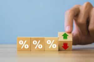 A hand flips a wooden block with a red down arrow and green up arrow, highlighting the effects of interest rates on mortgages, next to three blocks bearing percent signs on a table.