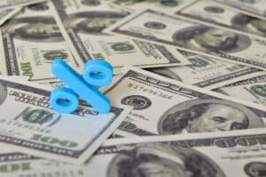 A blue percent symbol sits atop a scattered pile of 100 US dollar bills, visually representing the effects of interest rates on mortgages.