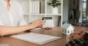 Person holding a calculator and pointing at it next to a clipboard with documents and a small model house on a desk, illustrating the effects of interest rates on mortgages.