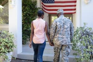 A woman and a man in military uniform hold hands while walking toward a house with an American flag hanging above the entrance, embodying the dream of homeownership and showing how VA Loans work for veterans.
