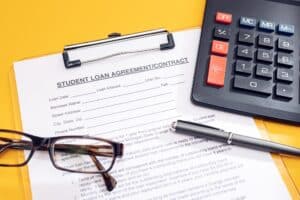 A student loan agreement form on a clipboard, with eyeglasses, a pen, and a calculator on a yellow background—highlighting the impact of student loans on mortgage approval.