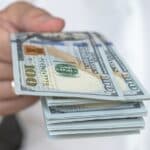 A person in a white shirt holds out a stack of U.S. hundred-dollar bills, symbolizing the cash flexibility often discussed in Hard Money vs Traditional Loans.