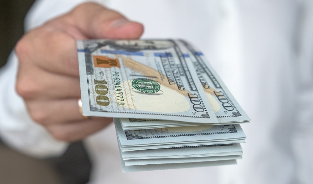 A person in a white shirt holds out a stack of U.S. hundred-dollar bills, symbolizing the cash flexibility often discussed in Hard Money vs Traditional Loans.