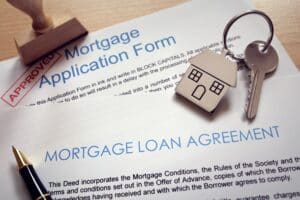 A mortgage application form and loan agreement are shown with a house-shaped keychain, keys, a stamp marked "approved," and a pen, highlighting the impact of student loans on mortgage approval.