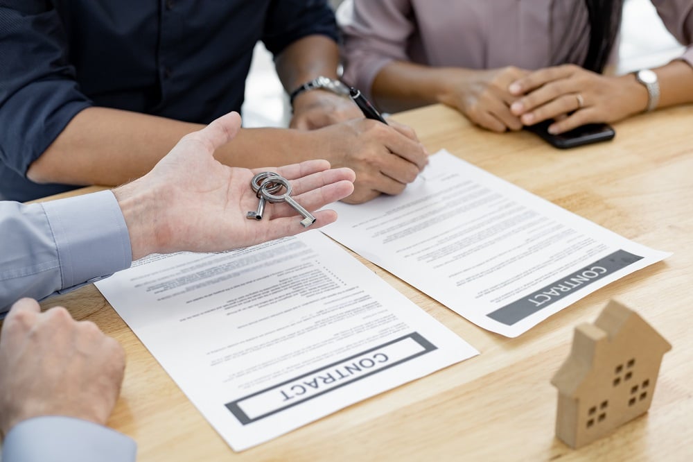 A person hands over house keys while two people, one acting as a Mortgage Co-Signer, sign real estate contracts on a wooden table, with a small wooden house model nearby.