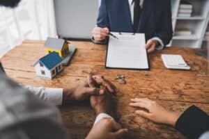 A couple sits at a table holding hands while a person in a suit shows them a contract; a model house, keys, and documents about a Mortgage Co-Signer are on the table.
