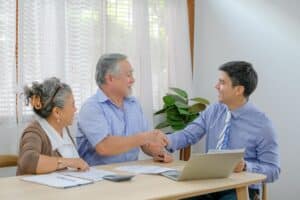 An elderly couple sits at a table with paperwork about Conventional Loans for Second Homes, while the man shakes hands with a younger professional who is using a laptop.