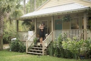 A person stands on the porch of a yellow house, hugging a child, with a dog beside them and greenery surrounding the porch—an inviting scene that’s perfect for those considering conventional loans for second homes.