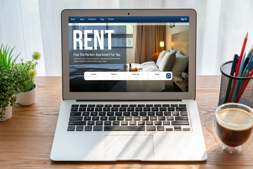 A laptop on a desk displays an apartment rental website with a search bar and a photo of a modern bedroom, hinting at investment opportunities like those supported by a DSCR loan. A plant, pens, and a cup of coffee are also on the desk.