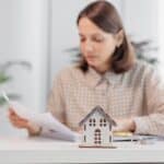 A woman sits at a desk reviewing documents, with a small model house and keys in the foreground—illustrating the careful steps involved in the Bridge Loan Refinancing Timeline.