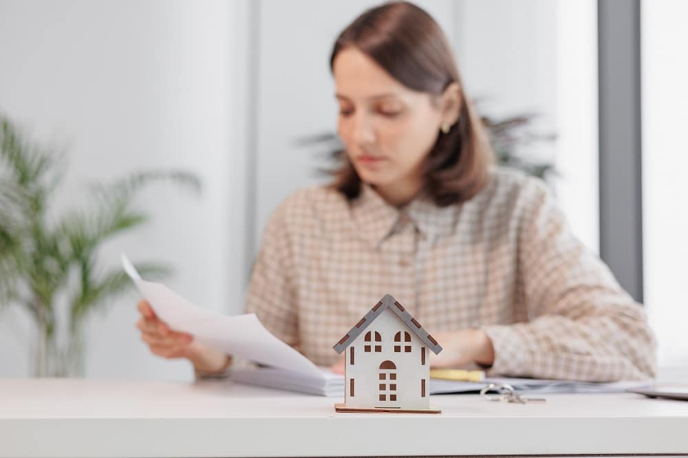A woman sits at a desk reviewing documents, with a small model house and keys in the foreground—illustrating the careful steps involved in the Bridge Loan Refinancing Timeline.