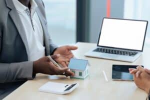 Two people sit at a desk with a laptop, tablet, calculator, and a small model house, discussing real estate or mortgage options and reviewing the Bridge Loan Refinancing Timeline.