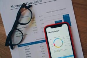 A pair of glasses, a mortgage calculator printout, and a smartphone displaying a monthly budget chart are placed on a wooden surface.