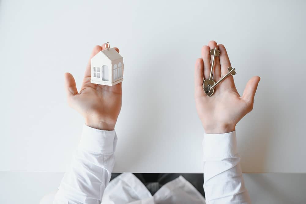 Person holding a small house model in one hand and a set of keys in the other, both hands visible on a white background.